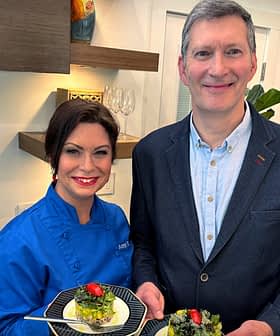 Two individuals, one in a blue chef jacket and the other in a blazer, holding plates of food in a kitchen. - Olive Oil Times