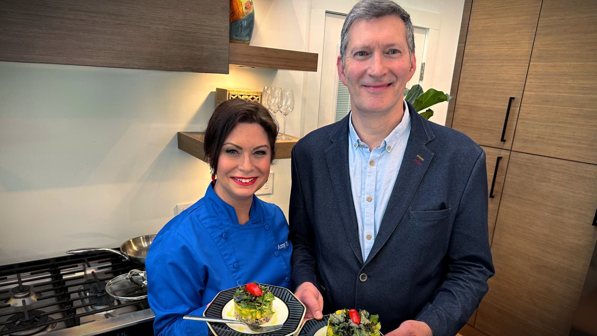 Two individuals, one in a blue chef jacket and the other in a blazer, holding plates of food in a kitchen. - Olive Oil Times