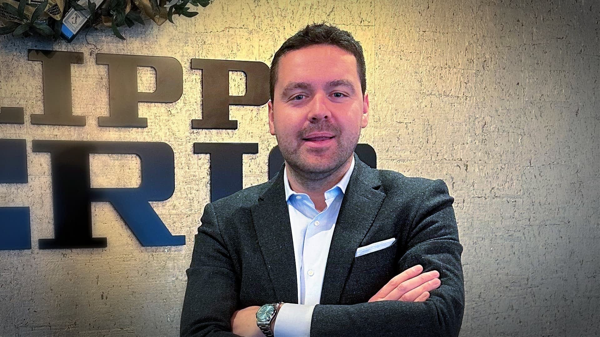 Man in a dark blazer and white shirt standing with arms crossed in front of a textured wall. - Olive Oil Times