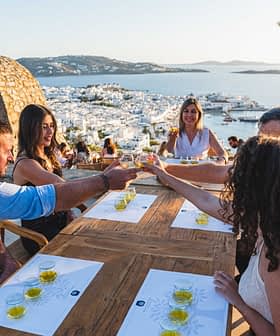A group of five people toasting with drinks at a wooden table overlooking a coastal view. - Olive Oil Times